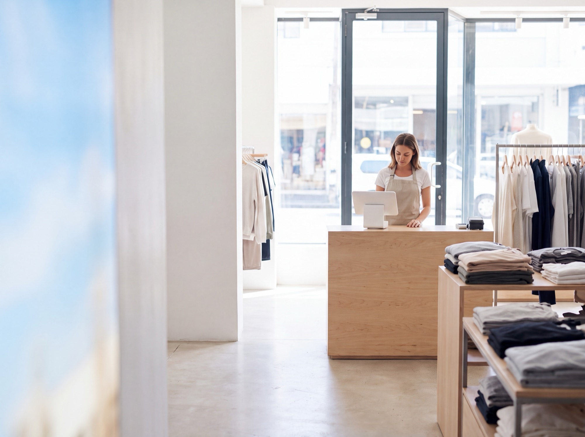 The interior of a bright modern shop: a sales assistant at a timber counter with a tablet terminal, rails of folded clothing and a daylit glass shopfront behind.