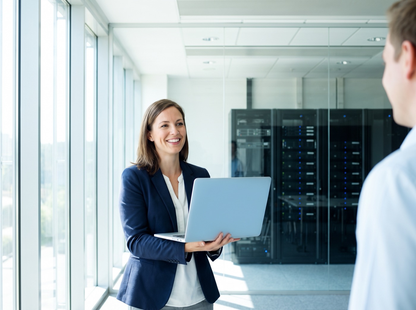 A consultant holding a laptop, smiling at a colleague in a bright office with server racks behind glass.